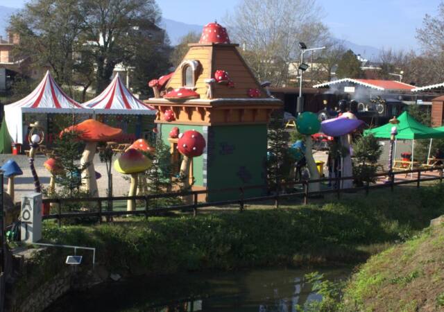 Η εικόνα παρουσιάζει το σκηνικό από τον Μύλο των Ξωτικών του 2022 στα Τρίκαλα. Στο κέντρο της εικόνας, υπάρχει ένα μικρό σπίτι με μανιτάρια στη στέγη και γύρω από αυτό. Δίπλα στο σπίτι, βλέπουμε πολύχρωμα μεγάλα μανιτάρια διακοσμητικά. Στο παρασκήνιο, υπάρχουν δύο σκηνές τσίρκου με ρίγες κόκκινες και λευκές. Αριστερά, υπάρχει μια τσουλήθρα και μερικοί επισκέπτες που περιηγούνται στο χώρο. Στα δεξιά, υπάρχουν πράσινες τέντες και μερικά ξύλινα κτίρια. Ο χώρος είναι πράσινος με δέντρα και θάμνους, και στο προσκήνιο υπάρχει ένα μικρό ρυάκι. Στο βάθος, μπορούμε να δούμε κτίρια και δέντρα, ενώ στο πίσω μέρος διακρίνονται βουνά.