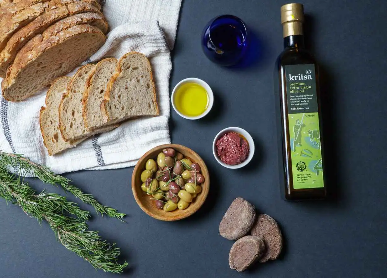 Το ελαιόλαδο ΠΓΕ Κριτσάς πάνω σε τραπέζι με ελιές, πάστα ελιών και ψωμί